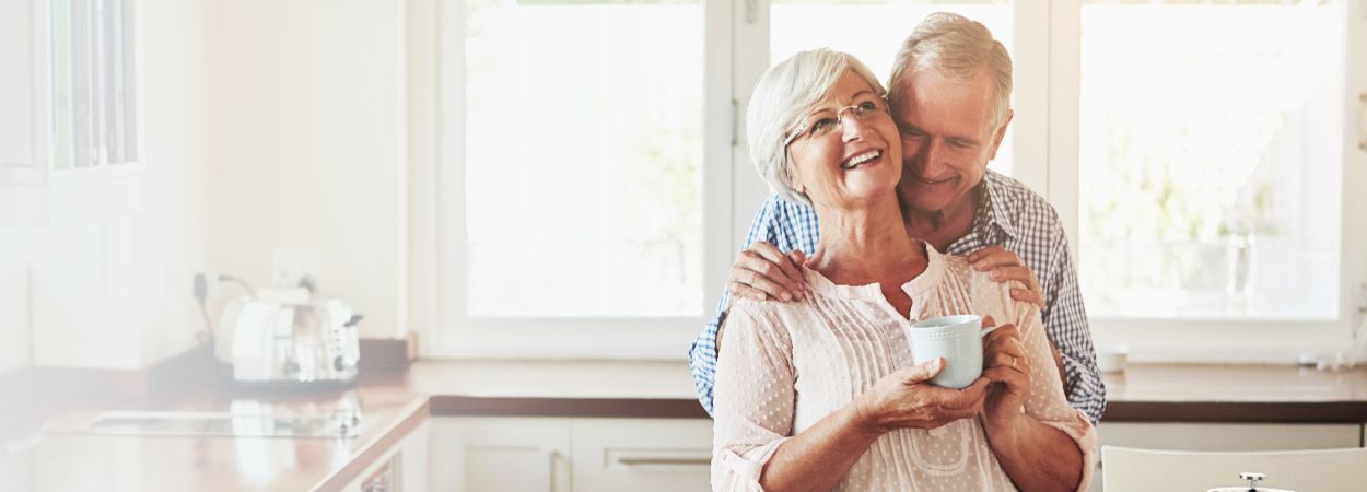 Smile hug senior couple with coffee kitchen love morning romance marriage home retirement happy man woman embrace support loyalty care with partner laughing banner (1)